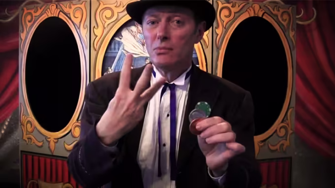 Charlie Frye presentando el efecto 3 Frye Coin con monedas mágicas en un fondo teatral