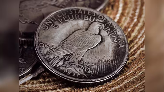 Reverso de una réplica del Dólar de la Paz de 1921, mostrando el águila en relieve sobre un fondo tejido, con otras monedas parcialmente visibles.