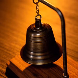 Spirit Bell by Tony Anverdi, campana metálica suspendida por cadena sobre base de madera con brazo curvo e iluminación cálida